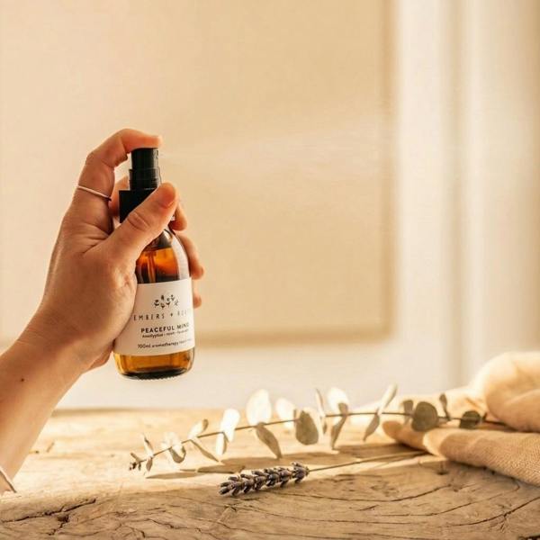 Hand spraying Peaceful Mind 100ml amber glass room mist by Abode Home & Gifts in warm sunlight over a wooden table with dried lavender and eucalyptus