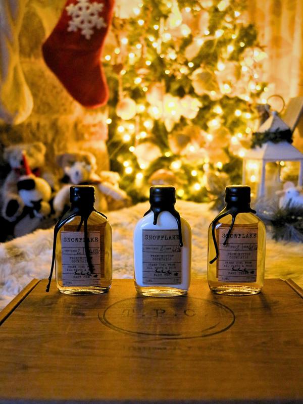 Three small glass bottles of Snowflake hand cream arranged on a wooden box in front of a warm, softly blurred Christmas tree and stockings.