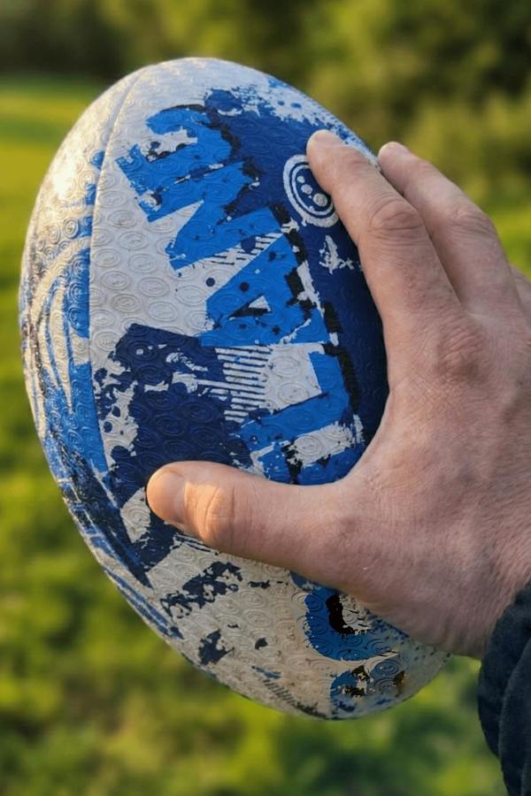 Hand holding a blue and white Optimum Scotland rugby ball outdoors with a grass background - Optimum
