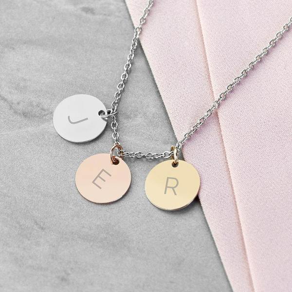 A personalised mixed-metal three-disc necklace featuring matte silver, rose gold, and yellow gold charms engraved with the uppercase initials 'J', 'E', and 'R' on a grey stone and pink fabric background.