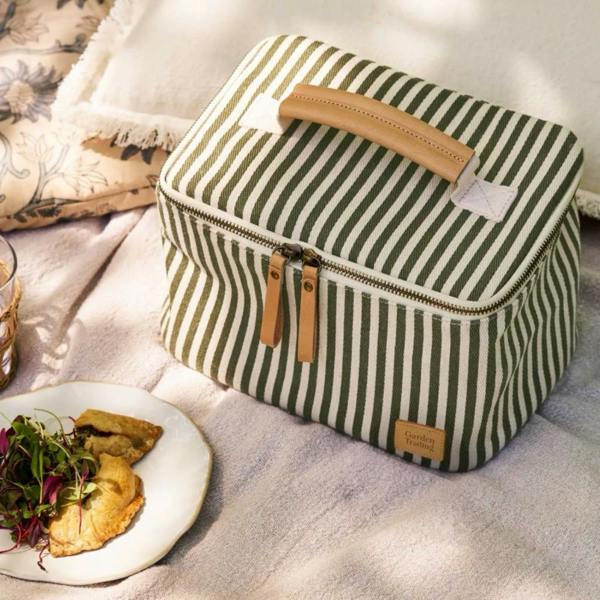 Moss green striped square lunch bag sitting on a picnic blanket next to a plate of food.