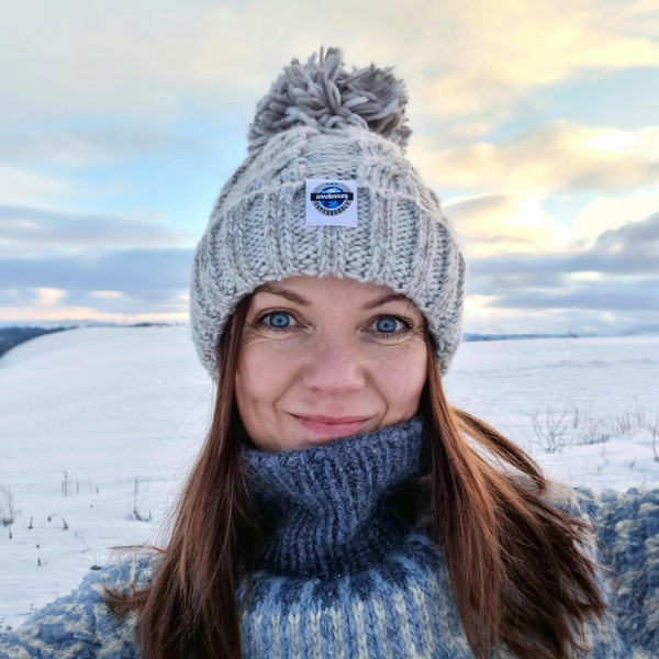 Woman standing with her dog, under a setting sun and milky blue skies is wearing a chunky cable knit silver grey Super Bobble hat. The hat has a large pom• pom, deep ribbed headband and contains reflective yarns and is lined with soft sherpa fleece.