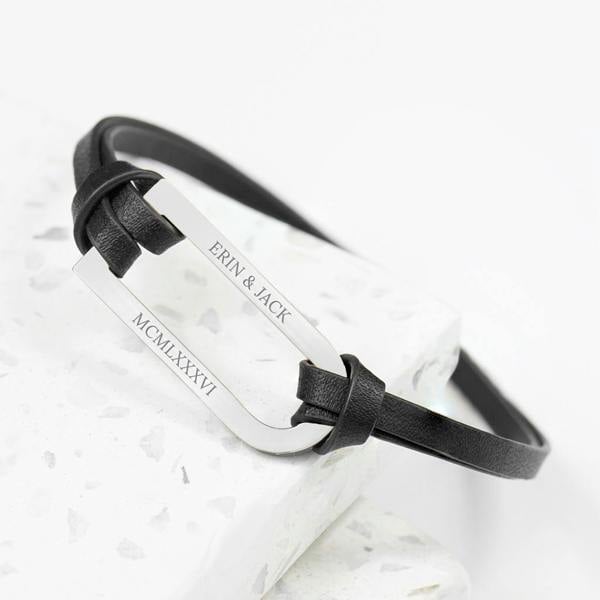 A custom engraved men's black leather bar bracelet from Abode Home & Gifts reading 'ERIN & JACK' and 'MCMLXXXVI' on a white marble display.