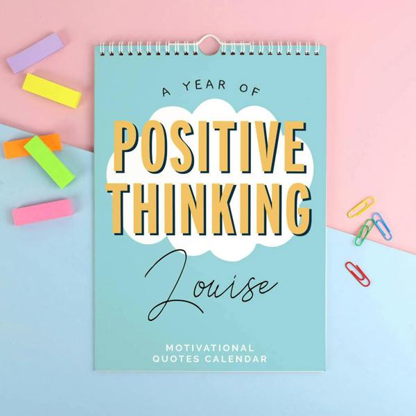 A light blue A4 wall calendar with a white cloud graphic in the centre. The text A YEAR OF POSITIVE THINKING is printed in yellow on the cloud, with the name Louise in script below it. The calendar is surrounded by colourful rectangular paper tags and paperclips on a pink and blue background.
