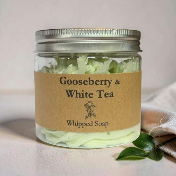A clear glass jar of Whipped Soap with a silver metal lid and a brown kraft paper label reading 'Gooseberry & White Tea'. The jar is filled with pale green whipped soap and sits on a light beige surface with a small green leaf prop.