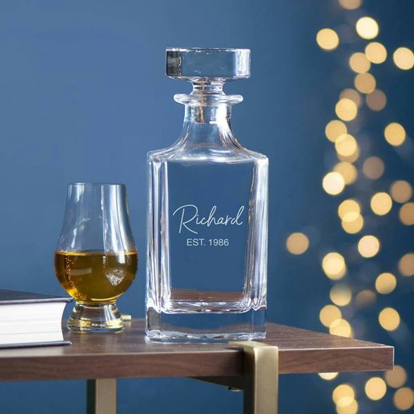 A premium clear glass square spirit decanter with a heavy round stopper, custom engraved with the elegant cursive script name 'Richard' above the block text 'EST. 1986', sitting on a wooden side table next to a filled tasting glass.