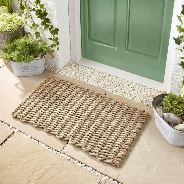 Woven doormat on a stone pathway leading to a green door with plants around.
