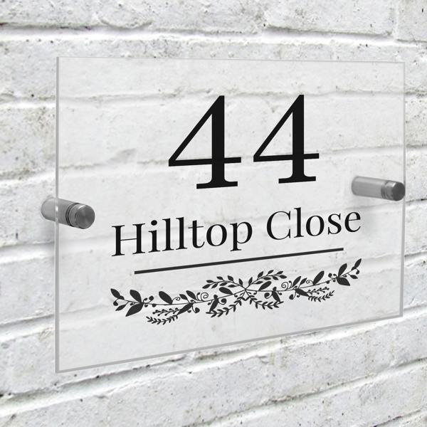 A clear rectangular acrylic house sign is mounted onto a white-painted brick wall using two silver metal stand-off fixings. The sign features a large black number '44' above the custom text 'Hilltop Close' and an elegant botanical leaf garland design.