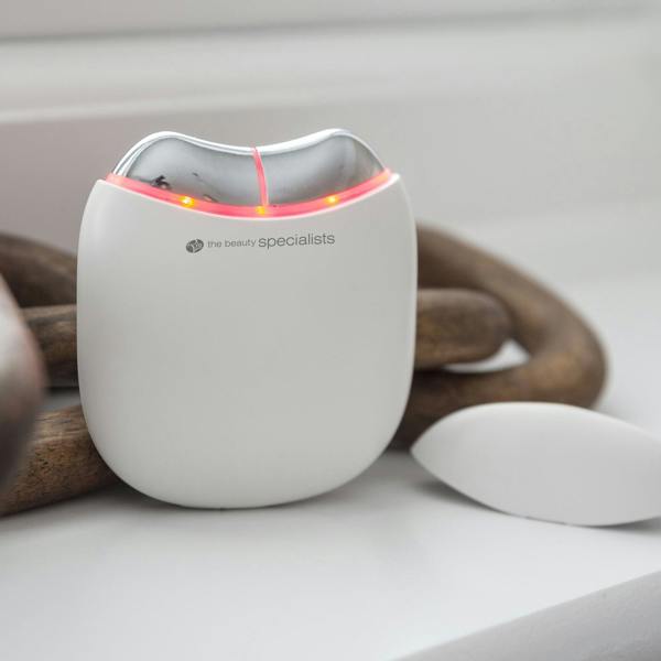A compact, white Rio Face Lift & Tone Facial Toner with red indicator lights for skin toning rests on a white surface beside a decorative wooden object. The device is branded the beauty specialists.
