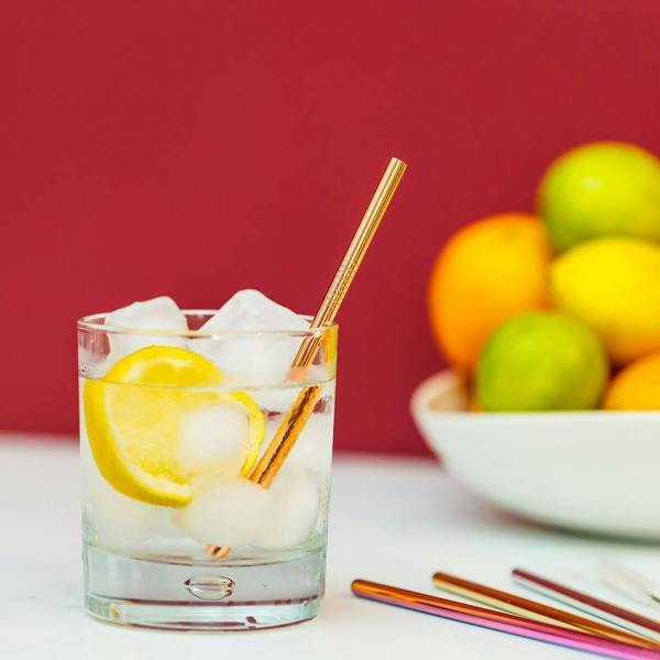 A short clear tumbler filled with ice, lemon, and a gold metal mini straw next to other multi-coloured straws.