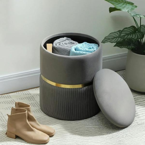 Grey storage ottoman with open lid holding folded blankets; round lid and gold-tone band