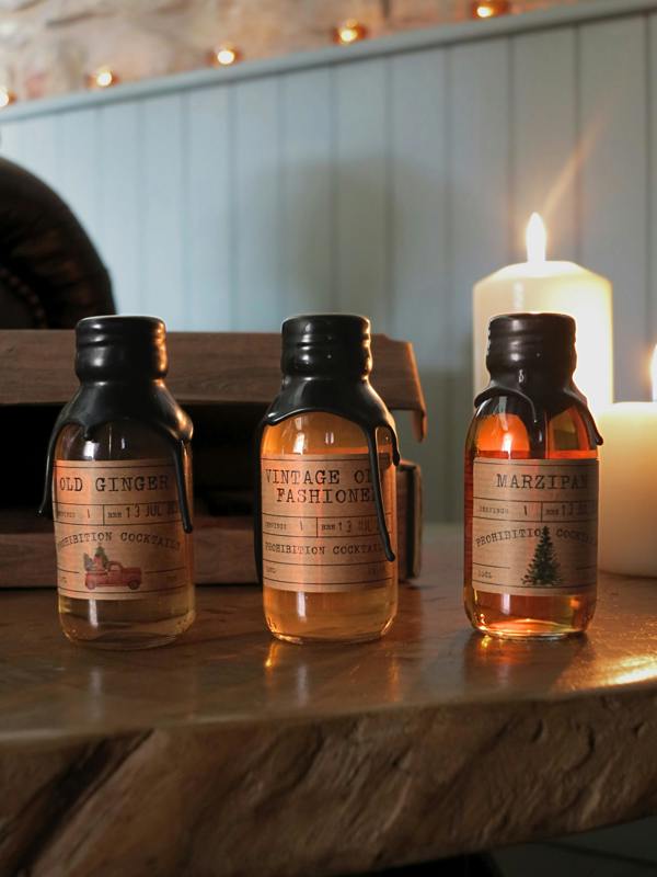 Three small amber glass bottles from Prohibition Cocktails with black caps and vintage-style labels sit on a wooden surface in a warmly lit room. A lit candle glows in the background against pale blue paneled walls.
