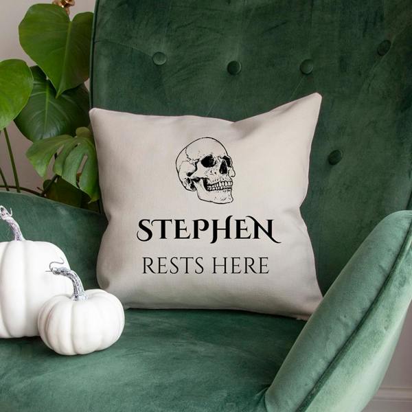 Square white cushion cover with skull graphic and Stephen Rests Here text resting on a dark green velvet armchair next to decorative white pumpkins