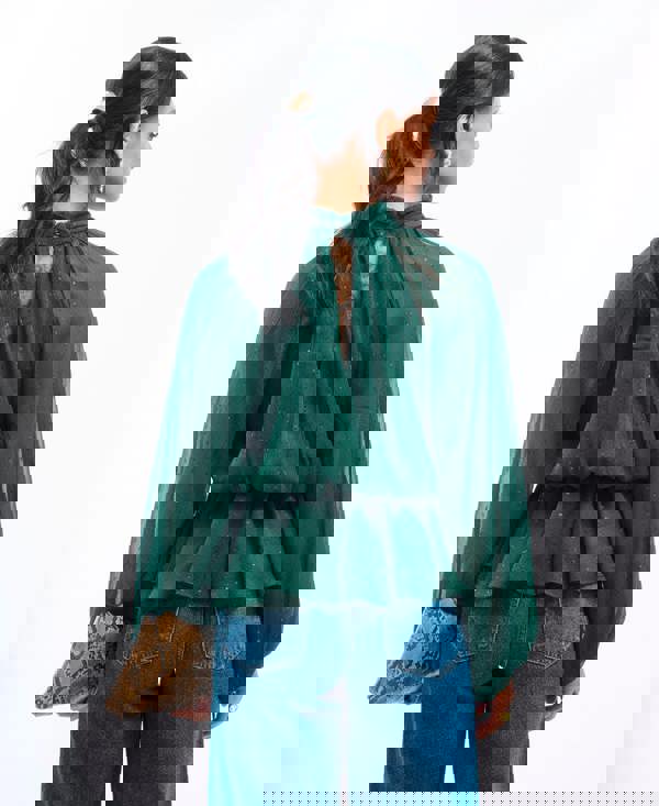 Woman wearing a green blouse with a sheer back and blue jeans, holding a clutch.