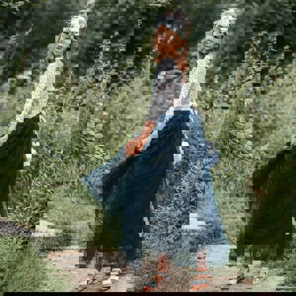 Young girl in a white lace top and blue skirt standing outdoors with greenery around.