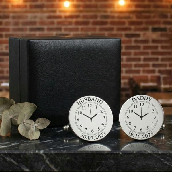 pair of silver-finished round cufflinks from Abode Home & Gifts engraved with Husband 26.07.2023 and Daddy 19.10.2025 alongside a textured black faux leather presentation case, resting on a polished dark marble base with a blurred background of warm string lights.