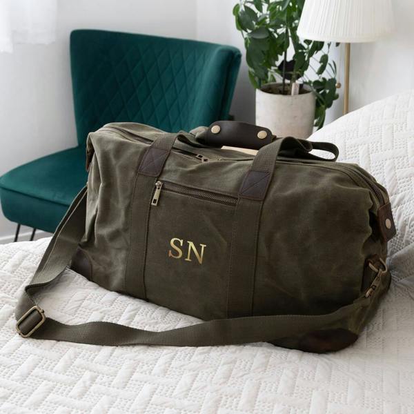 A personalised khaki waxed canvas holdall with gold initials SN on a white quilted bed runner.