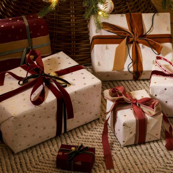 Selection of Christmas gifts beautifully wrapped in warm white handmade cotton paper with gold foil trees, stars, and berries, finished with velvet ribbons under a tree.