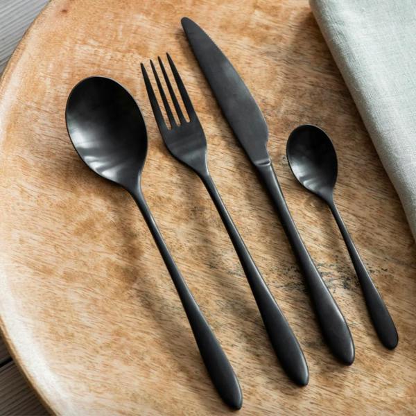 A 4-piece place setting of matte black stainless steel cutlery including a dinner knife, fork, dessert spoon, and teaspoon resting on a wooden charger plate.