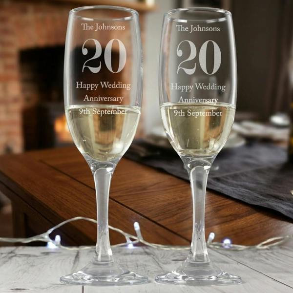 A pair of elegant personalised anniversary champagne flutes engraved with the name 'The Johnsons' and '20' filled with sparkling wine on a wooden dining table.