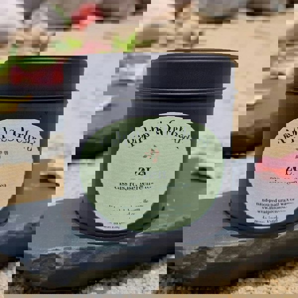 Candle tin labeled 'Ralph's Orchard Zen' on a stone surface with flowers in the background