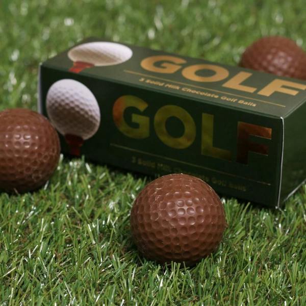 A gift box labeled 'GOLF' containing three solid milk chocolate golf balls, sitting on artificial grass next to loose chocolate golf balls.