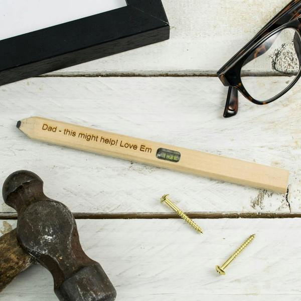 A custom laser-engraved wooden carpenter's pencil featuring a built-in spirit level reading Dad - this might help! Love Em, pictured on a rustic white table with a hammer, screws, and glasses.