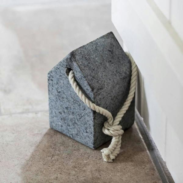 Minimalist house-shaped solid granite door stop with a knotted rope handle holding open a white interior door on stone flooring