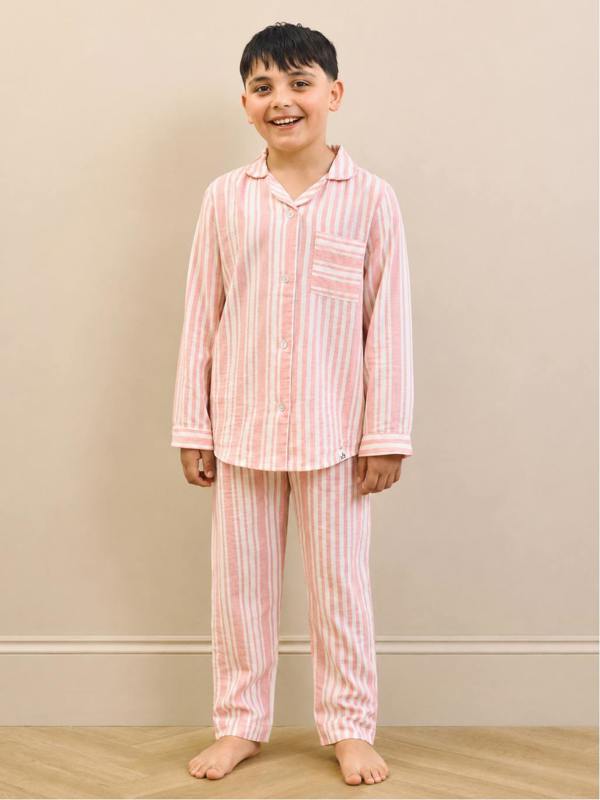 A young boy stands barefoot against a beige wall wearing a pink and white vertically striped pajama set with a long-sleeve button-up shirt and matching pants.