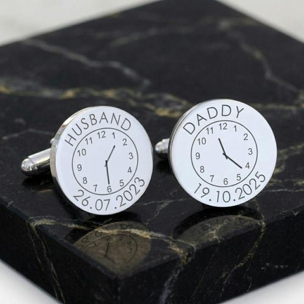 A pair of personalised silver finish round cufflinks engraved with Husband and Daddy alongside custom clock face time stamps, standing upright on a luxury square block of black marble with gold veins.