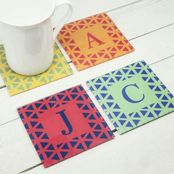 Set of four vibrant personalised glass coasters with custom initials A, J, and C on a white table