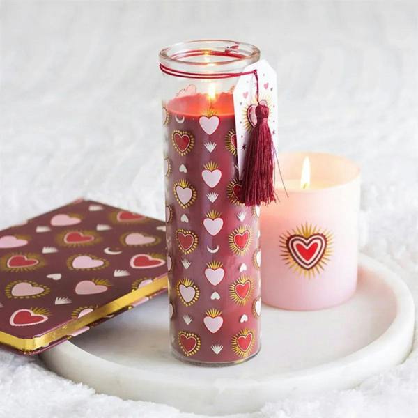 Printed tube candle in clear glass with pink heart pattern and red tassel, lit