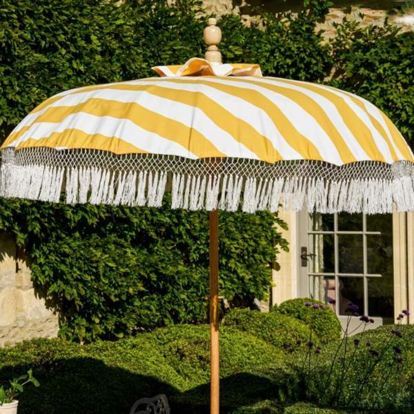 A luxury garden parasol with bright yellow and white canvas stripes and white cotton crochet fringe tassels. The umbrella has a central teak wood pole and stands on a manicured green lawn in front of a stone cottage wall draped in lush green ivy.