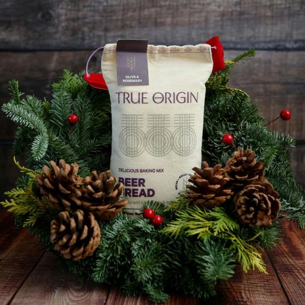 A fabric sack of Olive and Rosemary beer bread mix sitting in the centre of a festive green Christmas wreath decorated with pine cones and red berries on a wooden table.