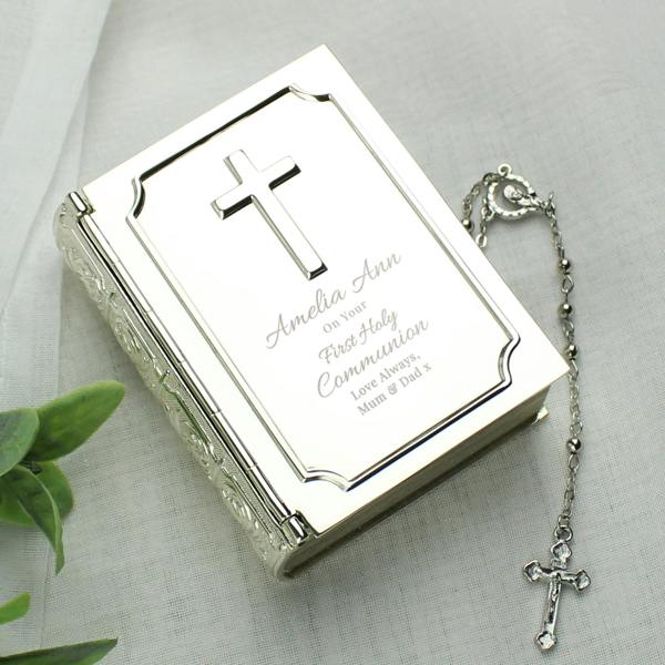 A top-down shot of a silver-finish bible trinket box by Abode Home & Gifts on a white fabric background, engraved with "Amelia Ann On Your First Holy Communion Love Always, Mum & Dad x" next to a silver crucifix rosary chain.