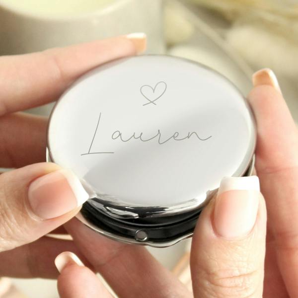 A close-up, high-angle shot of two hands with French-manicured nails holding a round, silver compact mirror. The mirror features a custom engraving of a heart icon and the name "Lauren" in a delicate cursive script, set against a blurred, elegant lifestyle background.