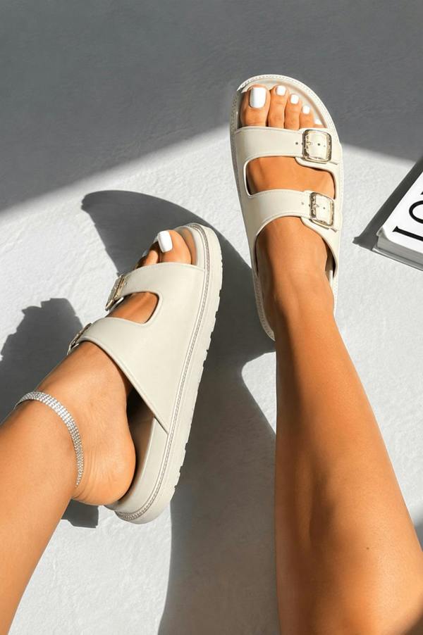 A pair of off-white double-strap platform slide sandals with silver buckle details, shown on a model's feet against a textured gray background with strong shadows.