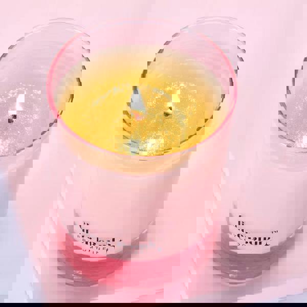 Pink candle labeled 'Glitter Bomb by Bloomtime' on a light pink background. The BLOOMTINE Glitter Bomb™ Gift Candle in a signature pink glass jar. This natural soy wax candle reveals a mesmerizing iridescent sparkle melt pool as it burns, releasing an addictive scent of creamy coconut, zesty lime, and vanilla sugar