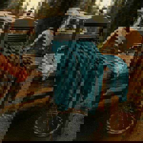 Manual hand wringer with a teal sweatshirt being squeezed, water dripping into a bucket, set in a forest campsite with a vintage van and tent in the background