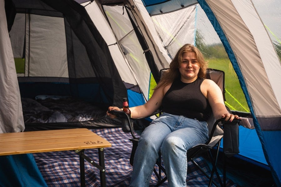 A woman sits inside the the OLPRO Cocoon V2 Poled Campervan Awning by OLPRO she is sat in an OLPRO camp chair next to a lightweight bamboo table