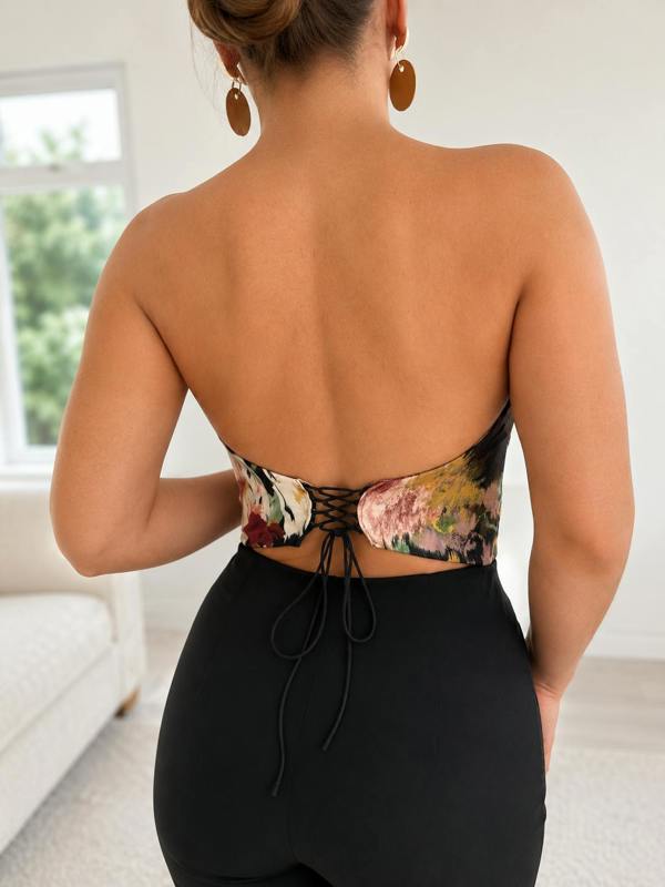 A person stands indoors with their back to the camera, wearing the Sarvin Ivy Corset Top—a waist-cinching floral halter style with lace-up detail—paired with high-waisted black pants. Their hair is up and they wear large round earrings.