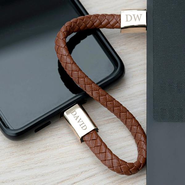 A braided tan leather men's charger bracelet unclasped and functioning as a USB charging cable connected between a black smartphone and a laptop.