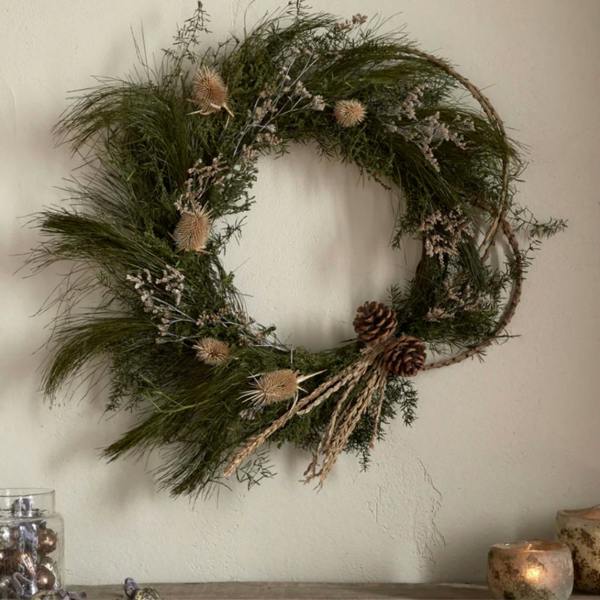 Elowen moss and thistle wreath hung on a neutral wall above a rustic wooden shelf.