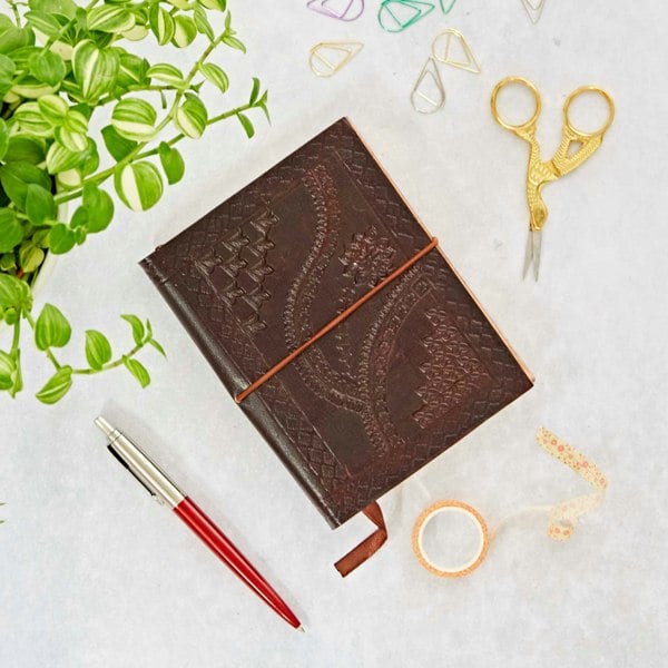 A Medium Brown Embossed Notebook, red pen, golden scissors, washi tape, and geometric paper clips are neatly arranged on a light surface with a green plant partially visible on the left.