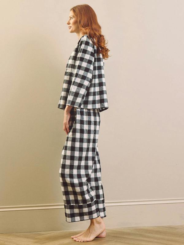 A woman with red hair stands in profile wearing a black and white gingham plaid boxy pyjama set, barefoot on a light wood floor against a beige wall.
