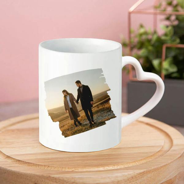 A white ceramic mug with a heart-shaped handle featuring a custom photo of a couple holding hands at sunset, displayed on a wooden board.
