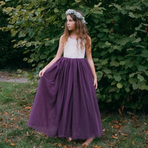 Young girl in a white top and purple skirt standing in front of green bushes.