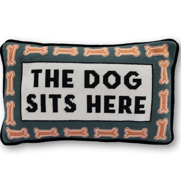 A rectangular hand-stitched needlepoint cushion with a muted teal background and orange dog bone borders, displaying the text "THE DOG SITS HERE" in black block letters inside a white central rectangle on a plain white background.