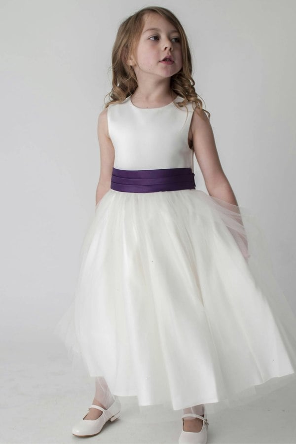 Young girl wearing an ivory dress with a purple belt on a plain background