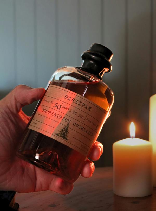 A hand holds a small amber glass bottle labeled 'MARZIPAN PROHIBITION COCKTAIL' in a warm, candlelit setting with a lit white pillar candle in the background.
